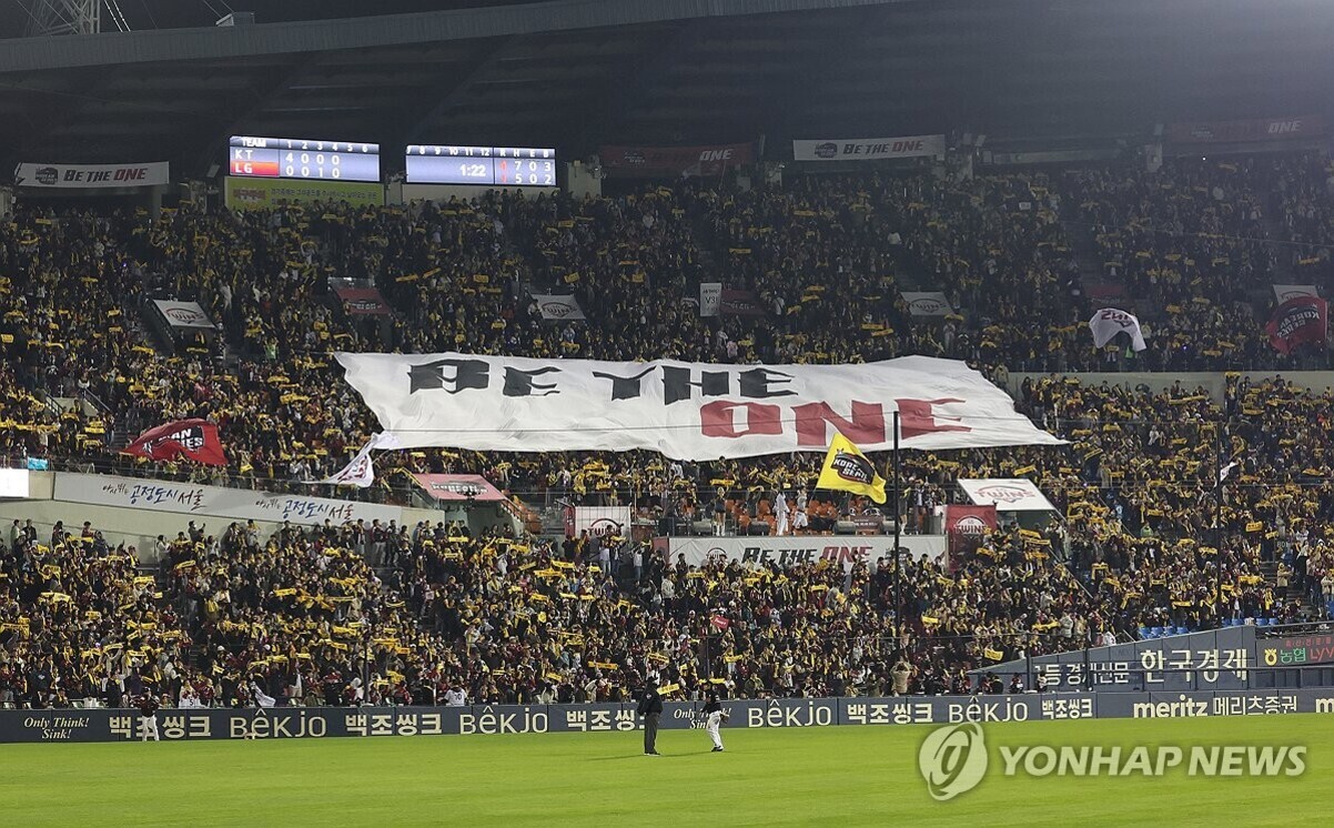 야구 경기장에서 관중들이 가득 찬 모습이며, 중앙에는 'BE THE ONE'이라고 적힌 대형 현수막이 펼쳐져 있다. 많은 사람들이 노란색 응원도구를 들고 응원하고 있다.