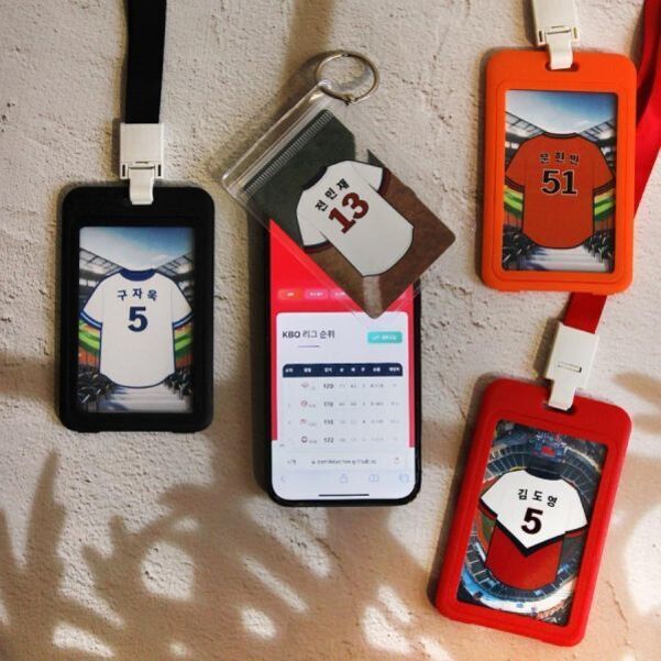 네 개의 명찰과 한 개의 키링이 벽에 걸려 있고, 각각 야구 유니폼 디자인과 이름 및 번호가 적혀 있다. 가운데에는 스마트폰이 놓여 있다.