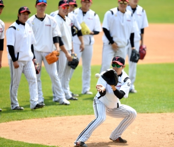 야구 유니폼을 입은 선수들이 야구장에서 연습을 하고 있으며, 한 명의 선수가 공을 잡으려고 글러브를 펼치고 있다.